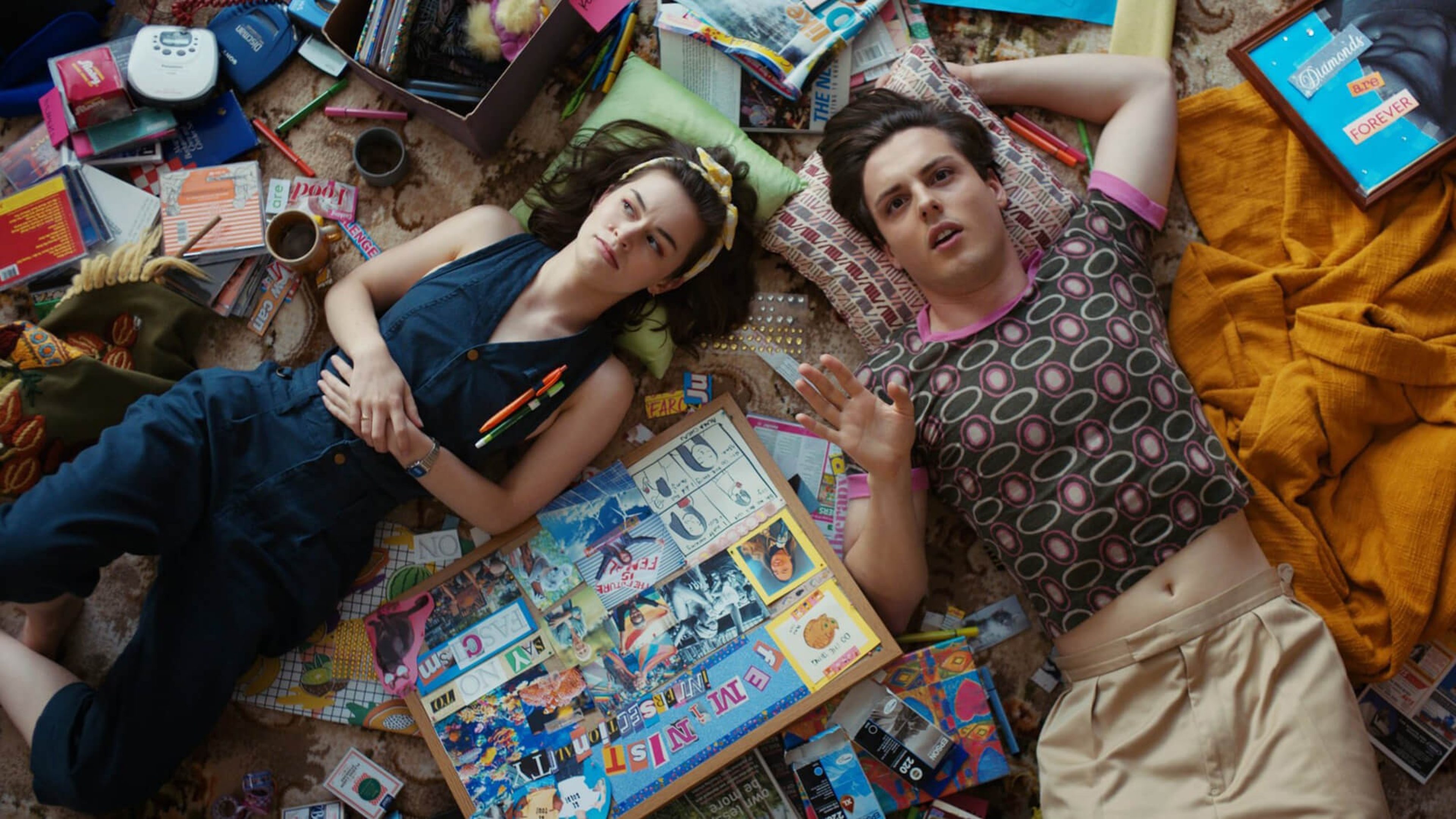 Zwei Jugendliche liegen auf dem Boden zwischen CDs, Büchern, Decken und Kissen