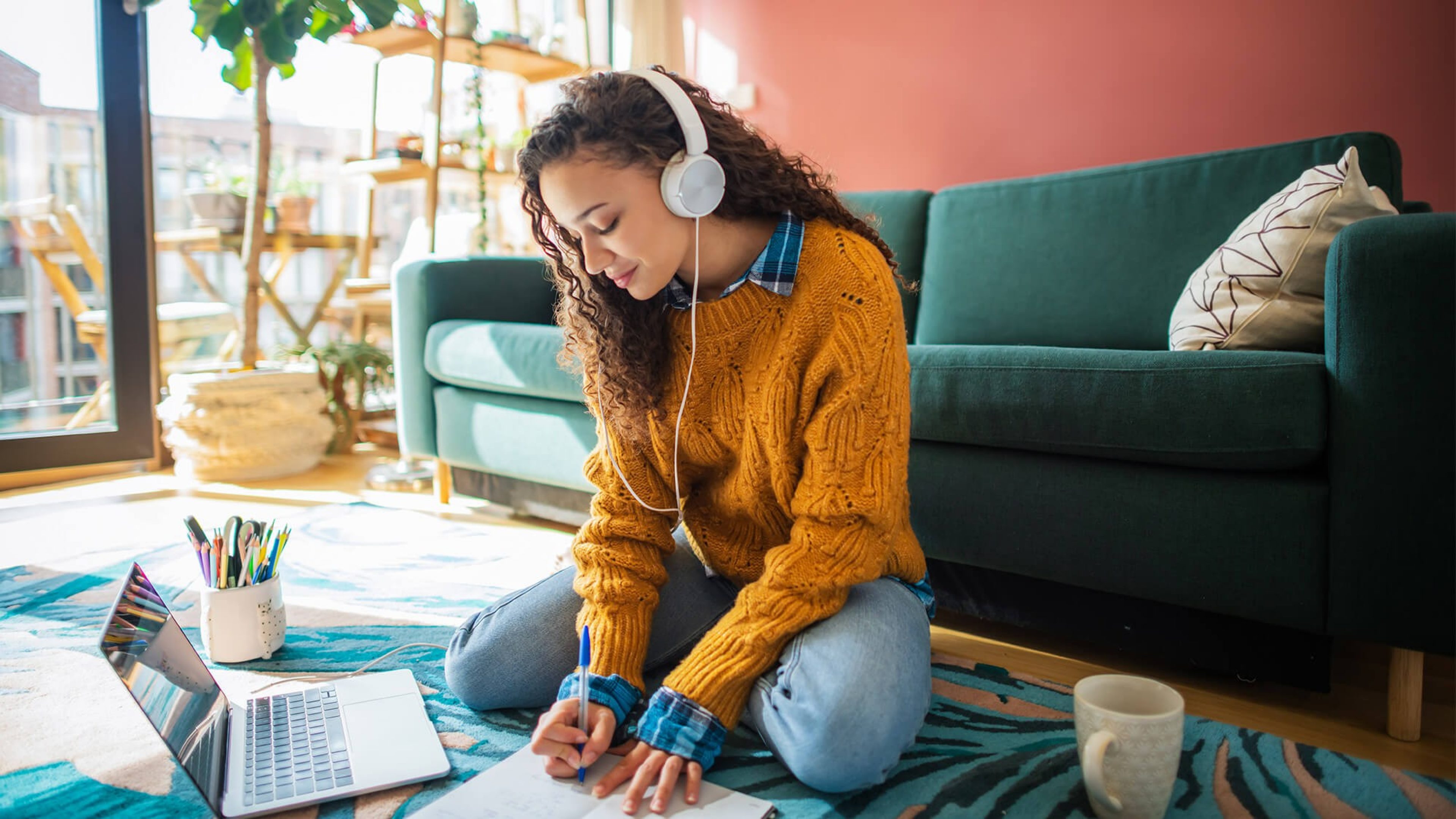 Junge Frau sitzt mit Kopfhörern auf dem Boden ihres Wohnzimmers vor einem Laptop und schreibt in ein Notizbuch