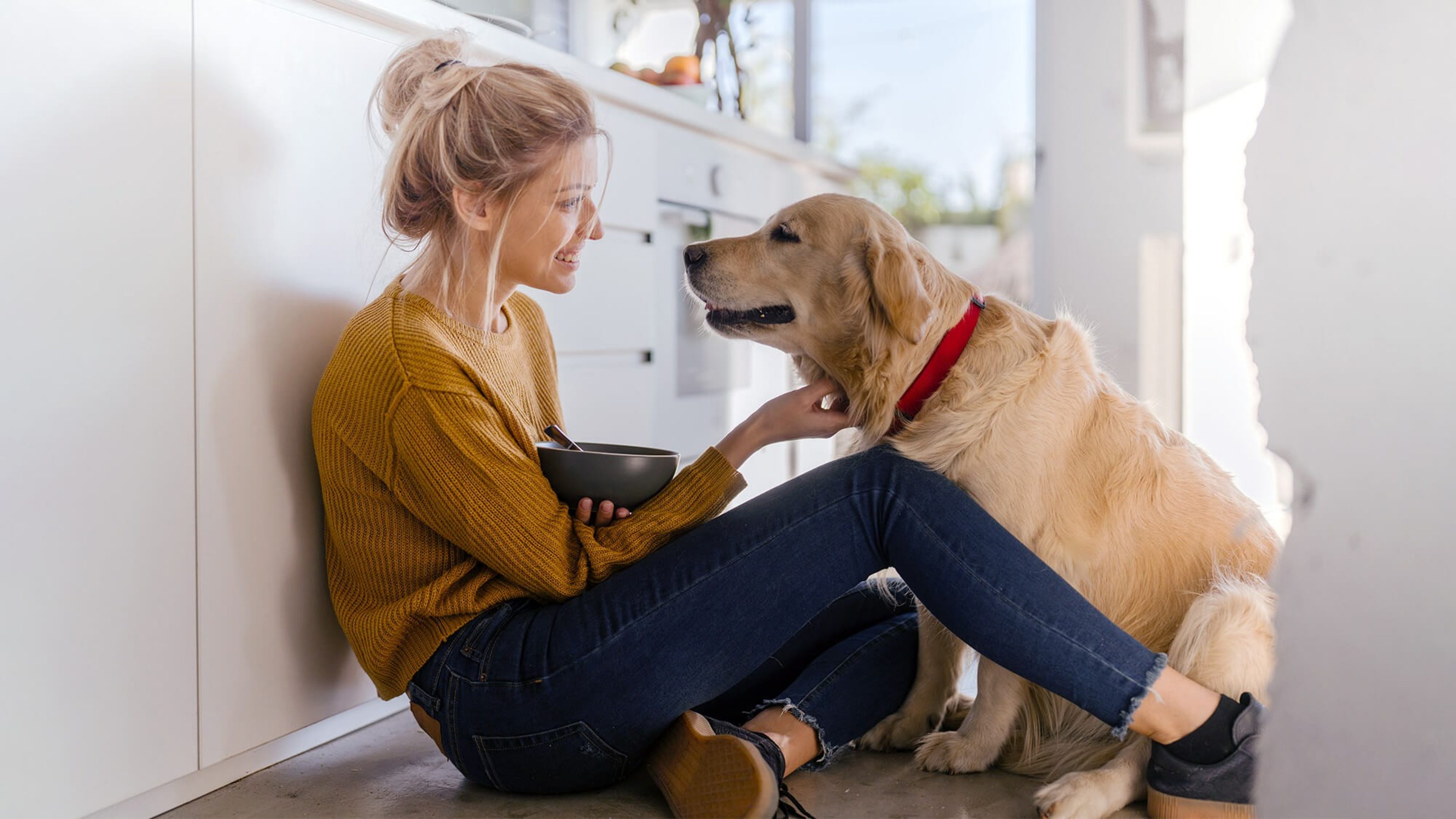 Frau sitzt auf dem Boden ihrer Küche und streichelt lächelnd ihren Hund, der vor ihr sitzt