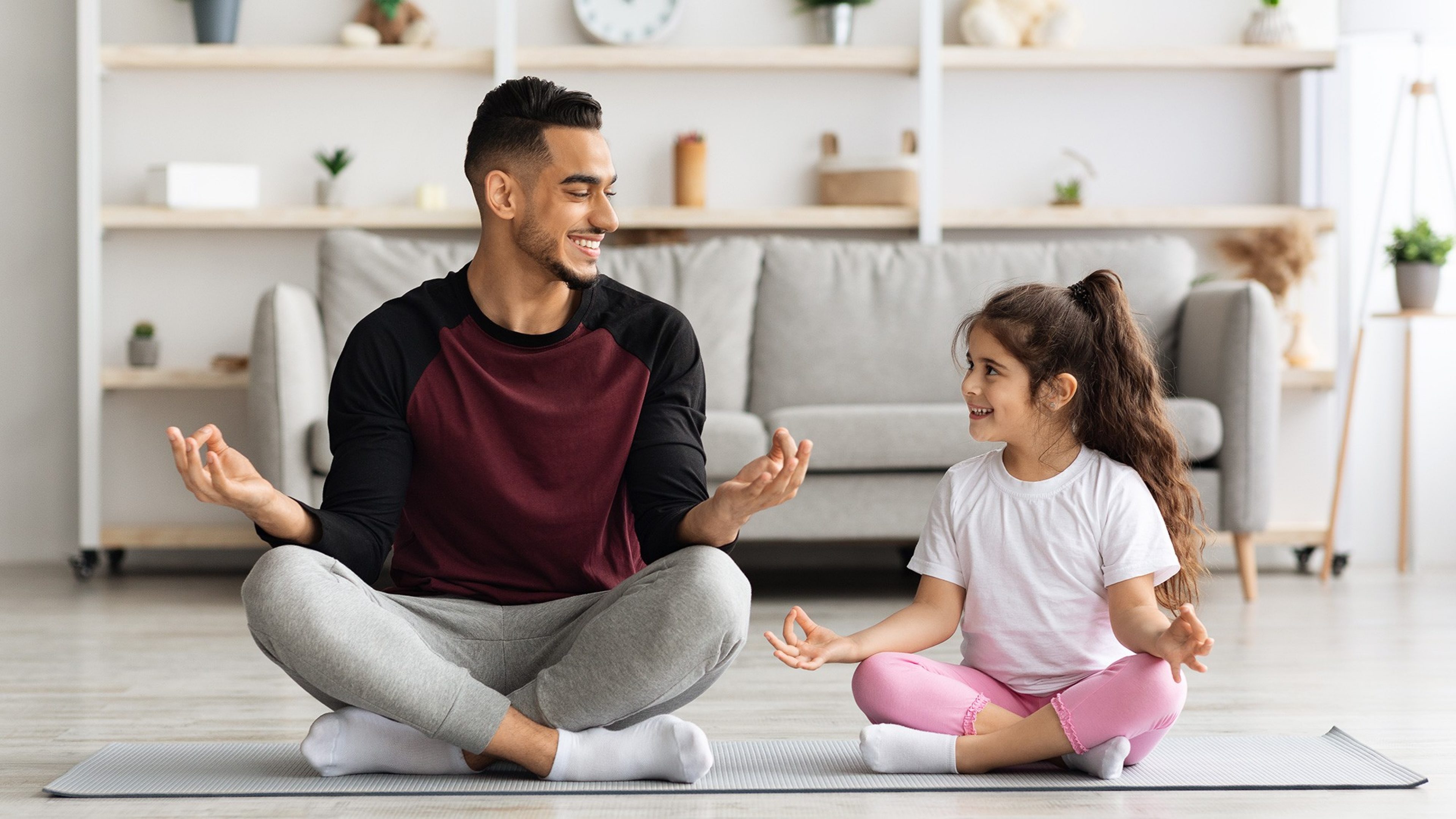 Vater und Tochter sitzen nebeneinander im Schneidersitz auf dem Boden eines hellen Zimmers und meditieren