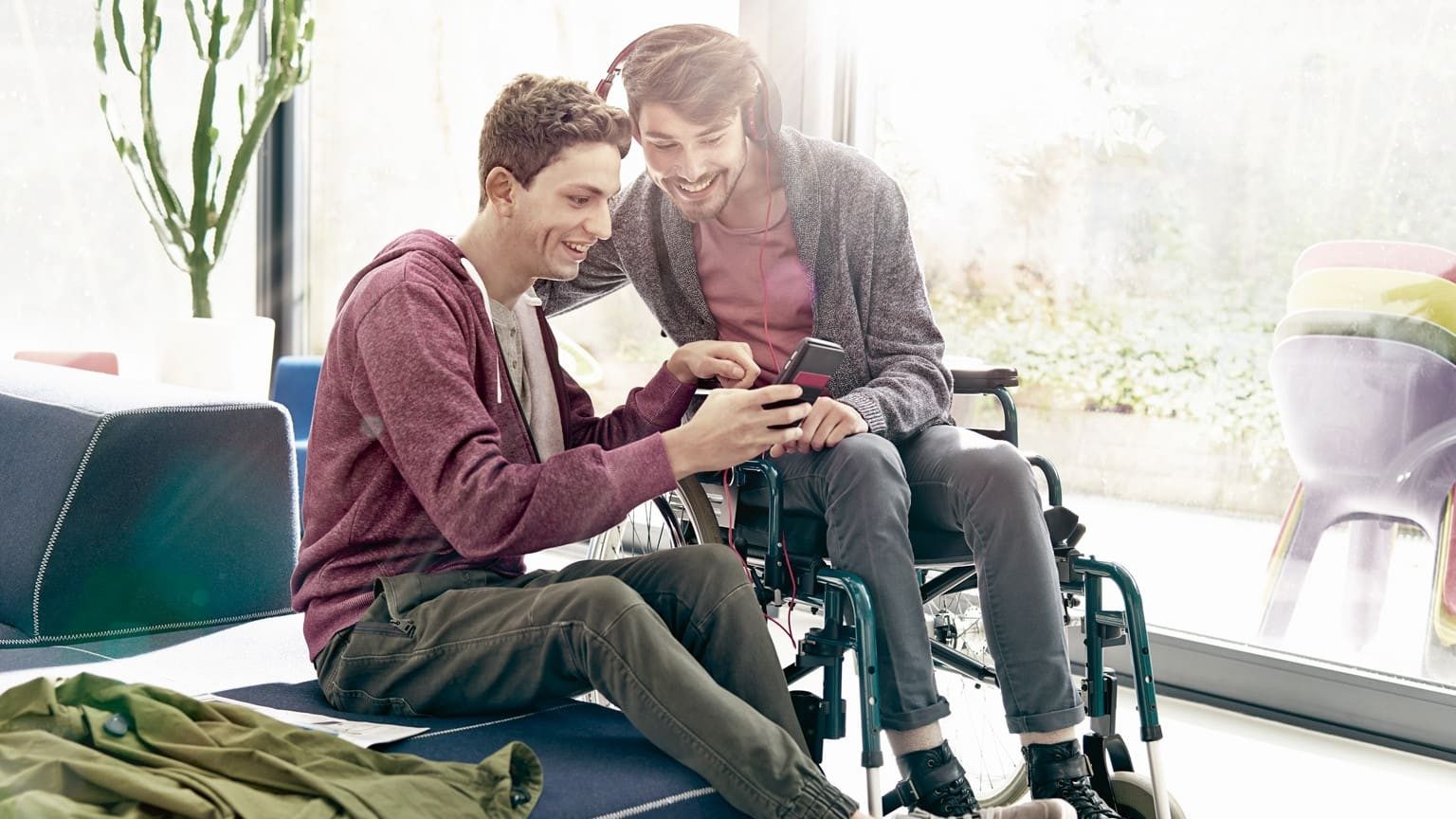 Junger Erwachsener im Rollstuhl und sieht sich im Kinderzimmer mit seinem Freund ein Video auf Handy an
