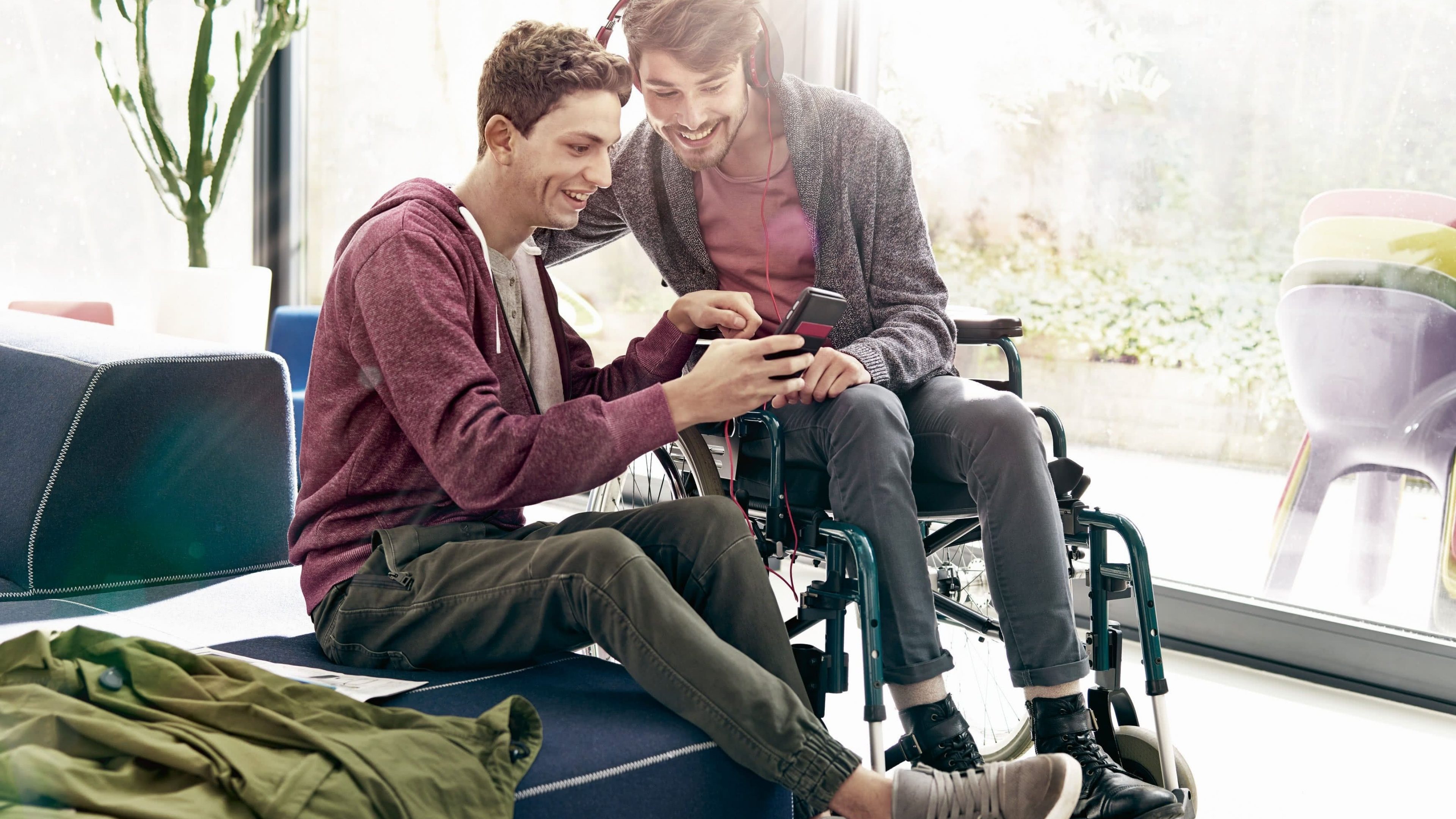 Junger Mann im Rollstuhl in einem Kinderzimmer blickt mit Freund auf ein Handy