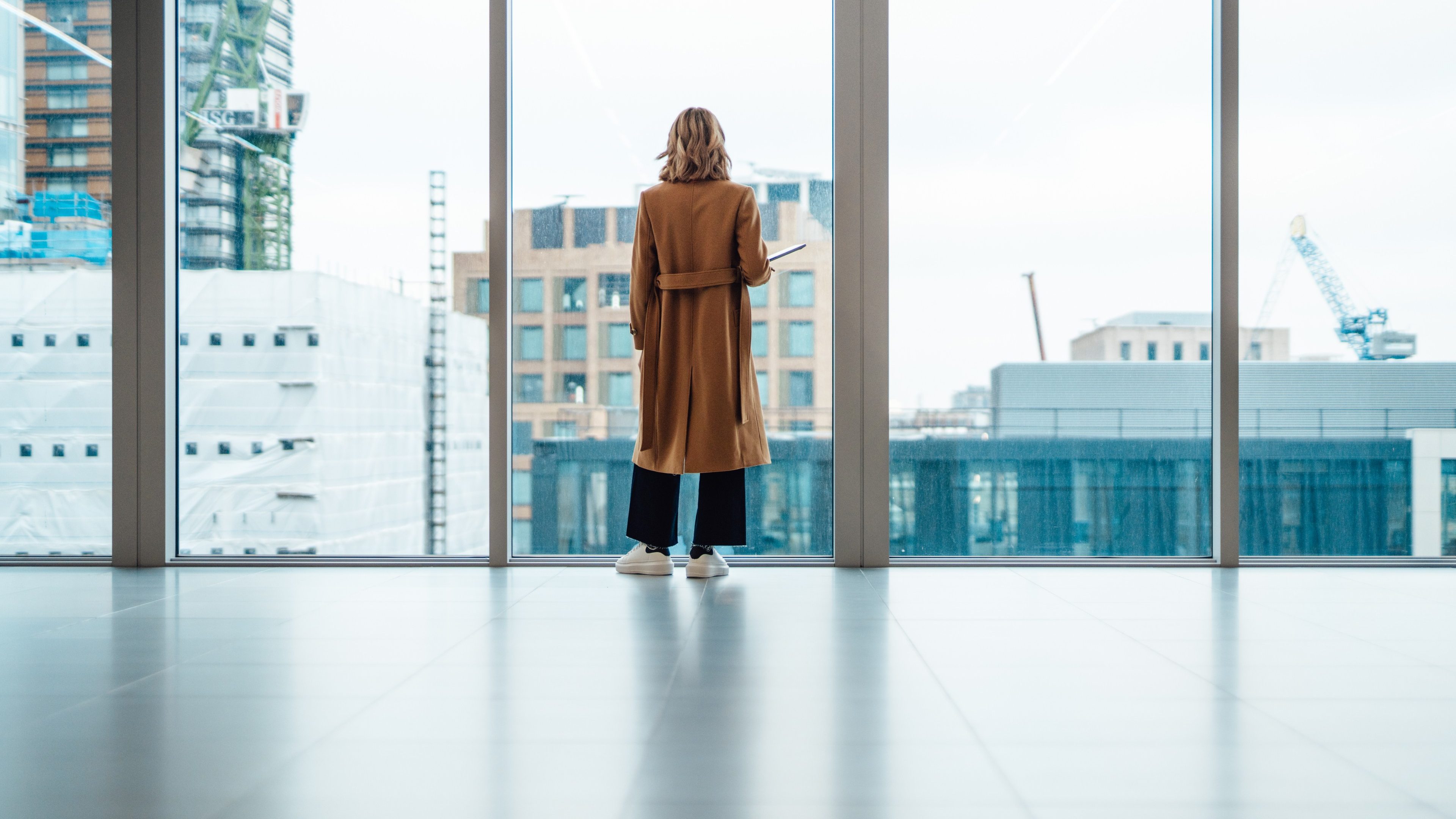 Back view of businesswoman standing and looking through window in conference room. She is thinking business strategy and making new business plan. Holding a laptop on her arm. Transgender female pursuing her dreams and successful career.